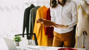 moda progettista l giovane asiatico donna Lavorando utilizzando computer portatile, tavoletta e sorridente mentre in piedi nel laboratorio rispondere su attività commerciale foto