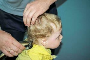 il primo taglio di capelli su il testa di un' poco ragazzo nel il suo vita. un' uomo è un' padre chi tagli il suo figli maschi capelli. radersi il riccioli via il del bambino testa con un elettrico rasoio foto