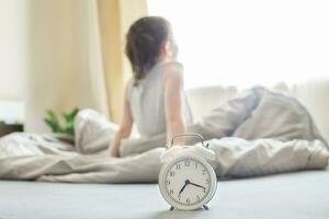 poco ragazzo seduta nel letto a casa nel il mattina su un' finestra sfondo con allarme orologio foto