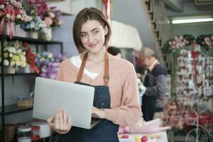 ritratto di uno giovane bianca femmina fioraio proprietario nel grembiule con il computer portatile nel luminosa e bellissimo fiore negozio memorizzare sorrisi con in linea shopping lavori, piccolo e-commerce attività commerciale, contento pmi imprenditore. foto