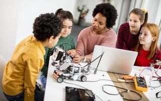 bambini felici con la loro insegnante di scienze afroamericana con laptop che programmano giocattoli elettrici e robot in classe di robotica foto