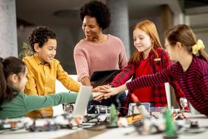 bambini felici con la loro insegnante di scienze afroamericana con laptop che programmano giocattoli elettrici e robot in classe di robotica foto