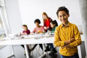 ragazzino carino in piedi di fronte a bambini che programmano giocattoli elettrici e robot in un'aula di robotica foto