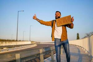 uomo è autostop su ciglio della strada provando per fermare macchina. lui è Tenere cartone con iscrizione. foto