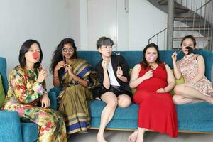giovane asiatico donna gruppo parlare cantare fabbricazione divertente beffardo facce espressione autoscatto ritratto carta oggetti di scena baffi cappello tubo naso capelli su blu vivente camera divano sedersi foto