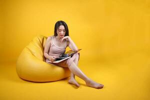 bellissimo giovane Sud est asiatico donna sedersi su un' giallo arancia beanbag posto a sedere colore sfondo rilassare leggere studia libro rivista foto