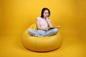 bellissimo giovane Sud est asiatico donna si siede su un' giallo beanbag posto a sedere arancia giallo colore sfondo posa moda stile elegante bellezza umore espressione riposo rilassare leggere libro pensare foto