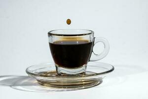 caffè gocciolina su trasparente bicchiere caffè espresso tazza piattino pieno di liquido caffè su bianca sfondo foto