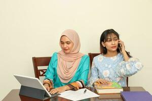 Due giovane asiatico malese musulmano donna indossare foulard a casa ufficio alunno seduta a tavolo Telefono computer libro documento studia parlare opera mescolarsi foto