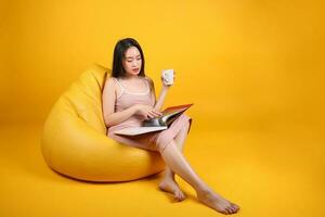 bellissimo giovane Sud est asiatico donna sedersi su un' giallo arancia beanbag posto a sedere colore sfondo rilassare leggere studia pensare libro rivista hold bianca caffè tazza foto