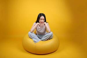 bellissimo giovane Sud est asiatico donna si siede su un' giallo beanbag posto a sedere arancia giallo colore sfondo posa moda stile elegante bellezza umore espressione riposo rilassare pensare foto