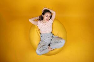 bellissimo giovane Sud est asiatico donna si siede su un' giallo beanbag posto a sedere arancia giallo colore sfondo posa moda stile elegante bellezza umore espressione riposo rilassare pensare emozione superiore Visualizza foto