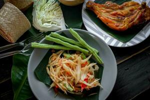 tailandese cibo piatto tutti e due nel Tailandia e Asia papaia insalata o come noi chiamata esso somtum è integrato con grigliato pollo e appiccicoso riso con fresco patatine fritte. servito su il nero di legno tavolo. foto