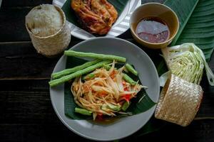 tailandese cibo piatto tutti e due nel Tailandia e Asia papaia insalata o come noi chiamata esso somtum è integrato con grigliato pollo e appiccicoso riso con fresco patatine fritte. servito su il nero di legno tavolo. foto