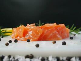 fresco crudo salmone trota filetto con cucinando ingrediente erbe aromatiche e Limone su bianca piatto al di sopra di nero sfondo, salmone trota fetta per fabbricazione sashimi o Sushi, salutare ricco omega nutrizione cibo aperitivo foto