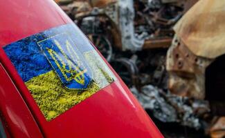 etichetta con ucraino bandiera e cappotto di braccia. vicino su giallo e blu etichetta con il ucraino bandiera su un' rosso auto contro il sfondo di rovine e bruciato macchine. Ucraina, kyiv - Maggio 06, 2023. foto