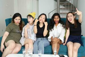 giovane asiatico donna gruppo parlare pettegolezzo Chiacchierare cantare divertente beffardo facce espressione su blu vivente camera divano foto