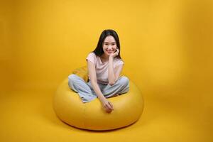 bellissimo giovane Sud est asiatico donna si siede su un' giallo beanbag posto a sedere arancia giallo colore sfondo posa moda stile elegante bellezza umore espressione riposo rilassare Sorridi foto