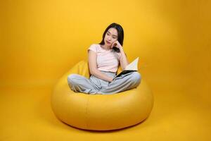 bellissimo giovane Sud est asiatico donna si siede su un' giallo beanbag posto a sedere arancia giallo colore sfondo posa moda stile elegante bellezza umore espressione riposo rilassare leggere libro pensare foto