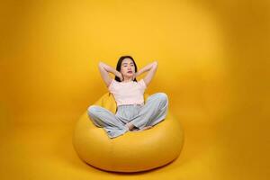 bellissimo giovane Sud est asiatico donna si siede su un' giallo beanbag posto a sedere arancia giallo colore sfondo posa moda stile elegante bellezza umore espressione riposo rilassare esercizio allungare yoga foto