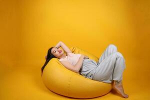 bellissimo giovane Sud est asiatico donna si siede su un' giallo beanbag posto a sedere arancia giallo colore sfondo posa moda stile elegante bellezza umore espressione riposo rilassare pensare emozione dormire foto