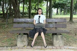 giovane asiatico Cinese donna all'aperto su parco panchina uso il computer portatile computer pensare leggere Guarda contento Meraviglia Sorridi Guarda davanti giù retrò foto