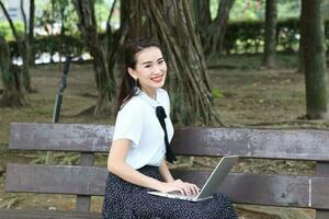 giovane asiatico Cinese donna all'aperto su parco panchina uso il computer portatile computer pensare leggere Guarda contento Meraviglia Sorridi Guarda inoltrare foto