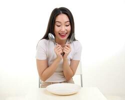 bellissimo giovane Sud est asiatico donna fare finta recitazione in posa Tenere vuoto forchetta cucchiaio nel mano mangiare gusto Guarda vedere bianca piatto su tavolo bianca sfondo Sorridi contento Uscito foto