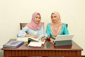 Due giovane asiatico malese musulmano donna indossare foulard a casa ufficio alunno seduta a tavolo parlare mescolarsi Guarda a computer libro documento studia discutere Sorridi contento foto