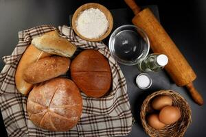 appena al forno pane pagnotta ciambella rotolo il giro lungo mescolare verità avvolto nel scacchi cucina tessuto tovagliolo asciugamano Grano fiore olio acqua sale uova rotolamento perno ardesia pietra al di sopra di nero sfondo foto