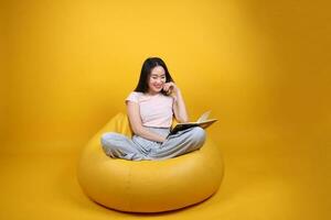 bellissimo giovane Sud est asiatico donna si siede su un' giallo beanbag posto a sedere arancia giallo colore sfondo posa moda stile elegante bellezza umore espressione riposo rilassare leggere libro pensare foto