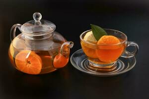 liquido tè Limone arancia fetta verde foglia cannella bastone nel trasparente bicchiere tazza di tè piattino teiera bollitore su nero sfondo foto