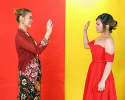 Due asiatico donna tradizionale kebaya e moderno vestito rosso giallo carta sfondo mani agitando Ciao saluti foto