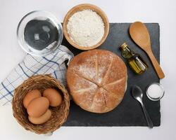 fresco al forno il giro pane pagnotta su nero ardesia pietra tavoletta con cucchiaio Grano fiore olio acqua sale cucina tovagliolo al di sopra di bianca sfondo foto