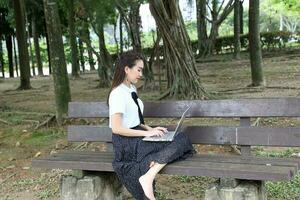 giovane asiatico Cinese donna all'aperto su parco panchina uso il computer portatile computer pensare leggere Guarda contento Meraviglia foto