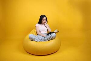 bellissimo giovane Sud est asiatico donna si siede su un' giallo beanbag posto a sedere arancia giallo colore sfondo posa moda stile elegante bellezza umore espressione riposo rilassare leggere libro pensare foto