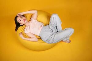 bellissimo giovane Sud est asiatico donna si siede su un' giallo beanbag posto a sedere arancia giallo colore sfondo posa moda stile elegante bellezza umore espressione riposo rilassare pensare emozione dormire superiore Visualizza foto