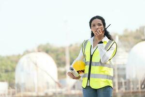 femmina ingegnere con elmetto protettivo con prodotto petrochimico fabbrica sfondo. asiatico donna Tenere tavoletta, Piano e walkie talkie. foto