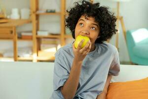 contento bella ragazza pungente verde Mela a casa. bellissimo africano americano giovane donna mangiare fresco frutta e sorridente. salutare cibo vegano vegetariano dieta concetto. salutare merenda pulito cibo. foto