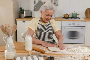 contento anziano donna cucinando nel cucina. elegante più vecchio maturo grigio dai capelli signora nonna impastare Impasto infornare biscotti. vecchio nonna cucinare fatti in casa cibo. domestico casalinga lavori di casa concetto. foto