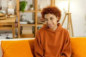 bellissimo africano americano ragazza con afro acconciatura sorridente seduta su divano a casa interno. giovane africano donna con Riccio capelli ridendo. la libertà felicità spensierato contento persone concetto. foto