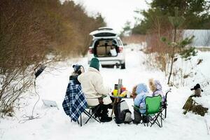 giovane donna con bambini nel inverno foresta su un' picnic. madre e tre bambini contro essi auto con Aperto tronco. foto