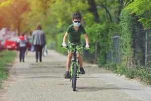 bambino felice che si diverte vicino a casa con una bicicletta in una bella giornata di primavera indossando una maschera protettiva per il virus pandemico covid19 del coronavirus