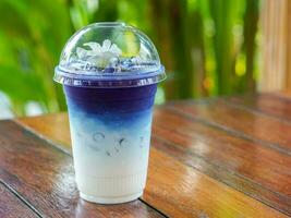 viola e blu farfalla pisello latte nel un' chiaro plastica bicchiere, posto su un' di legno tavolo, su sfocatura verde foglia sfondo, natura, cibo, bevanda foto