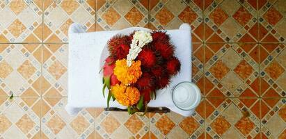 superiore Visualizza di fresco ghirlanda fiore con rambutan, Drago frutta e bicchiere di acqua su tavolo con colorato piastrella sfondo foto