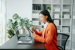 bellissimo asiatico donna utilizzando il computer portatile e tavoletta mentre seduta a sua Lavorando posto. concentrato a opera. foto