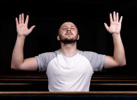 un uomo cristiano in camicia bianca è seduto con le mani in alto e prega con cuore umile nella chiesa