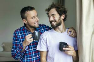 giovane gay coppia nel amore guardare su il finestra. Due giovane androgino uomini sorridente insieme e avendo caffè. foto