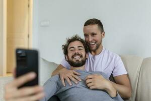 carino giovane gay coppia video chiamata loro amici nel loro vivente camera a casa. Due maschio Gli amanti sorridente allegramente mentre saluto loro amici su un' smartphone. giovane gay coppia seduta insieme. foto