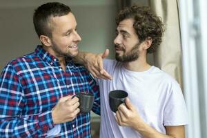 giovane gay coppia nel amore guardare su il finestra. Due giovane androgino uomini sorridente insieme e avendo caffè. foto